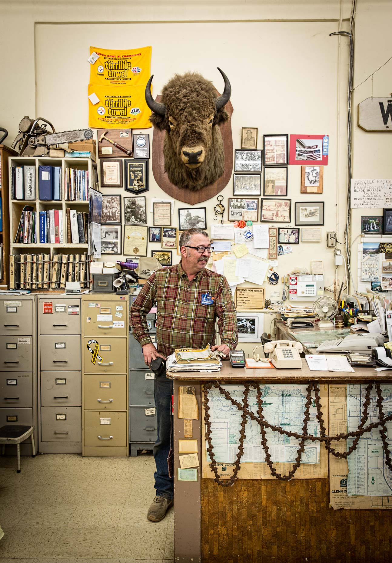 Portrait of man in cluttered hardware store under stuffed buffalo bison head on wall. Willows, California by David Zaitz