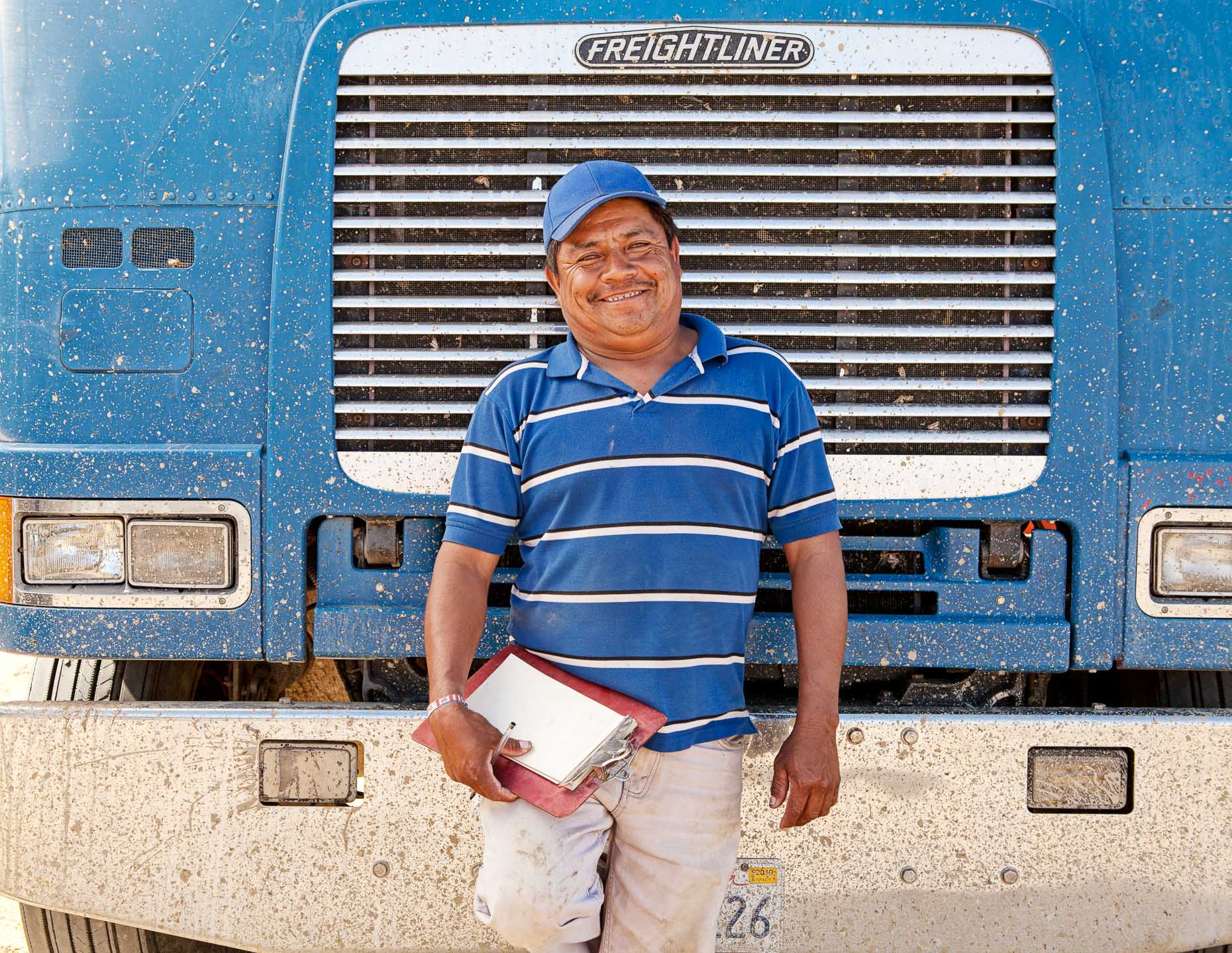 Truck driver with white and blue striped shirt standing in front of chrome grill of big rig truck by David Zaitz.