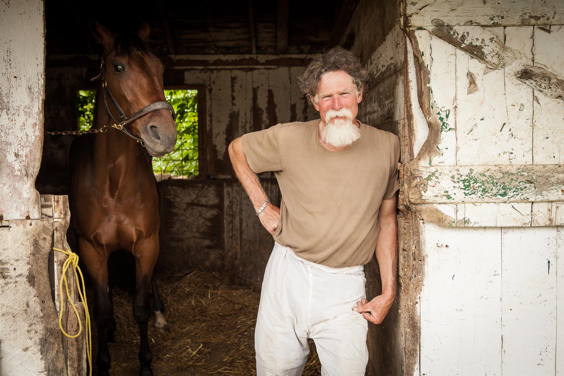Portrait of man with beard standing in horse stable with horse in background by David Zaitz