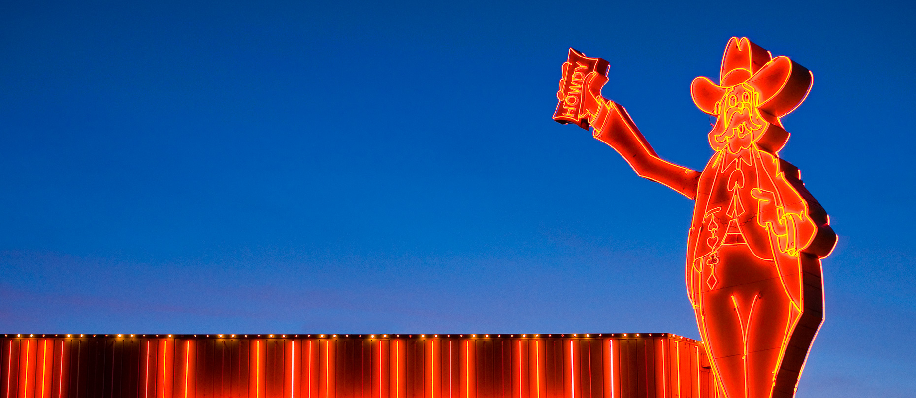Neon sign of man wearing hat holding money, sign says HOWDY by David Zaitz