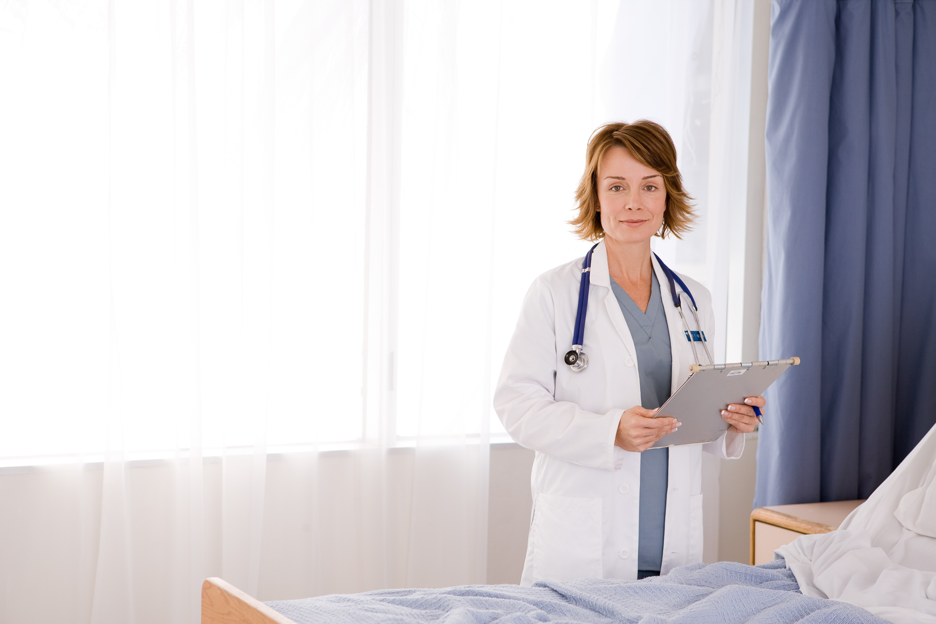 Portrait of medical doctor with clipboard next to hospital bed. Medical, healthcare. David Zaitz Photography.