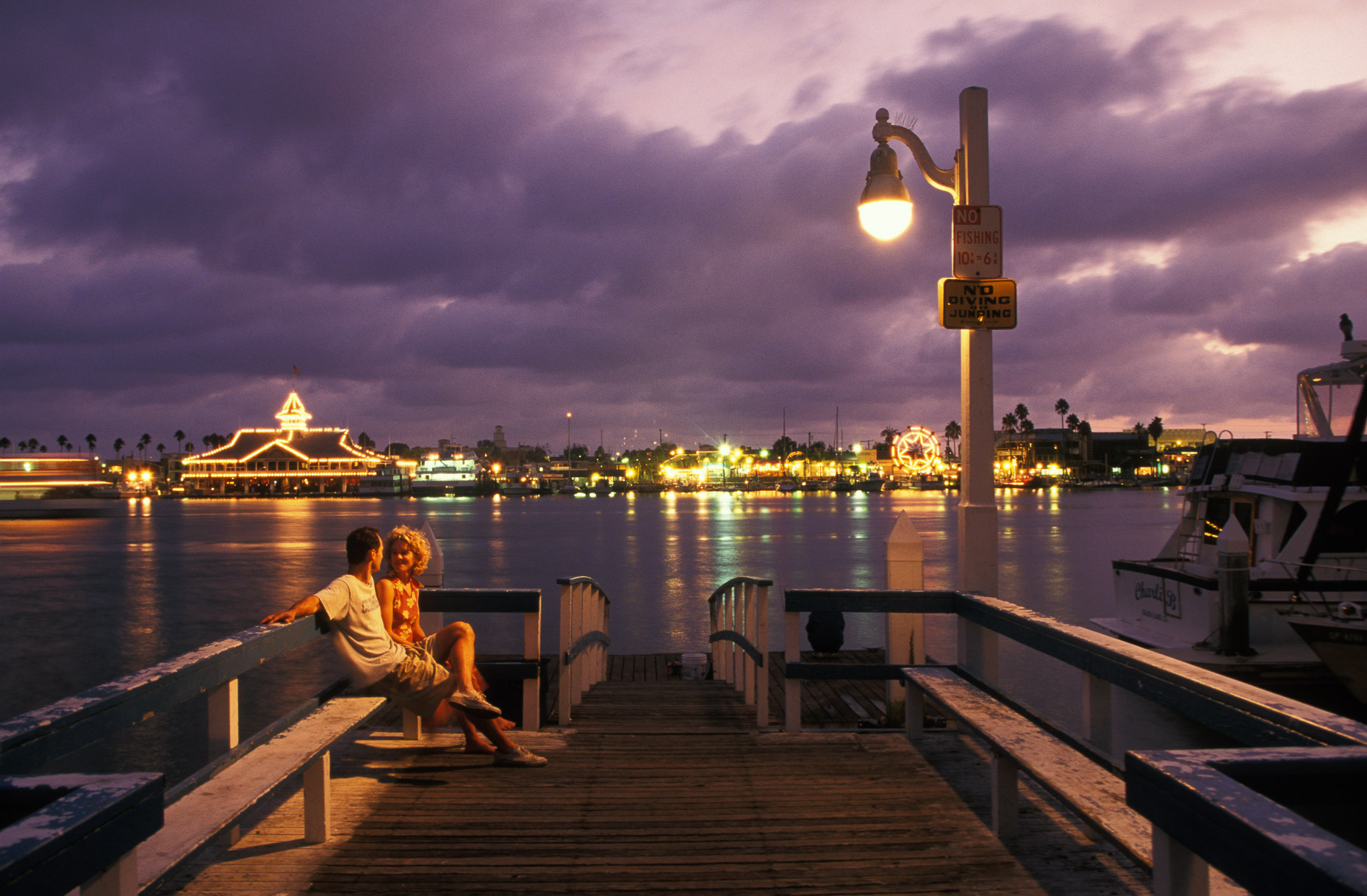 Man and woman sitting on bench on pier with sunset in background by David Zaitz.