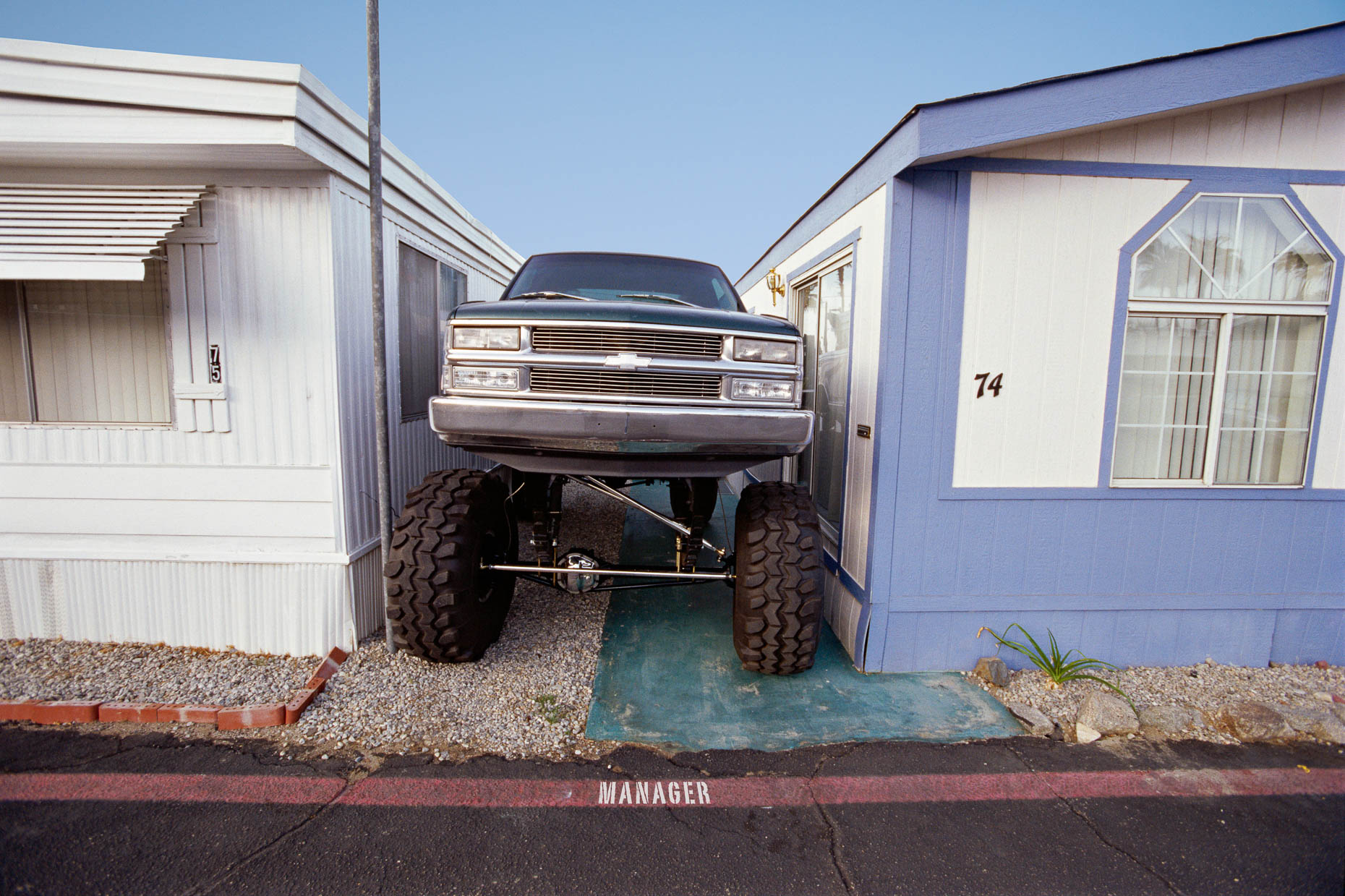 Large pickup truck parked between two mobile homes in a tight squeeze with MANAGER painted on pavement by David Zaitz