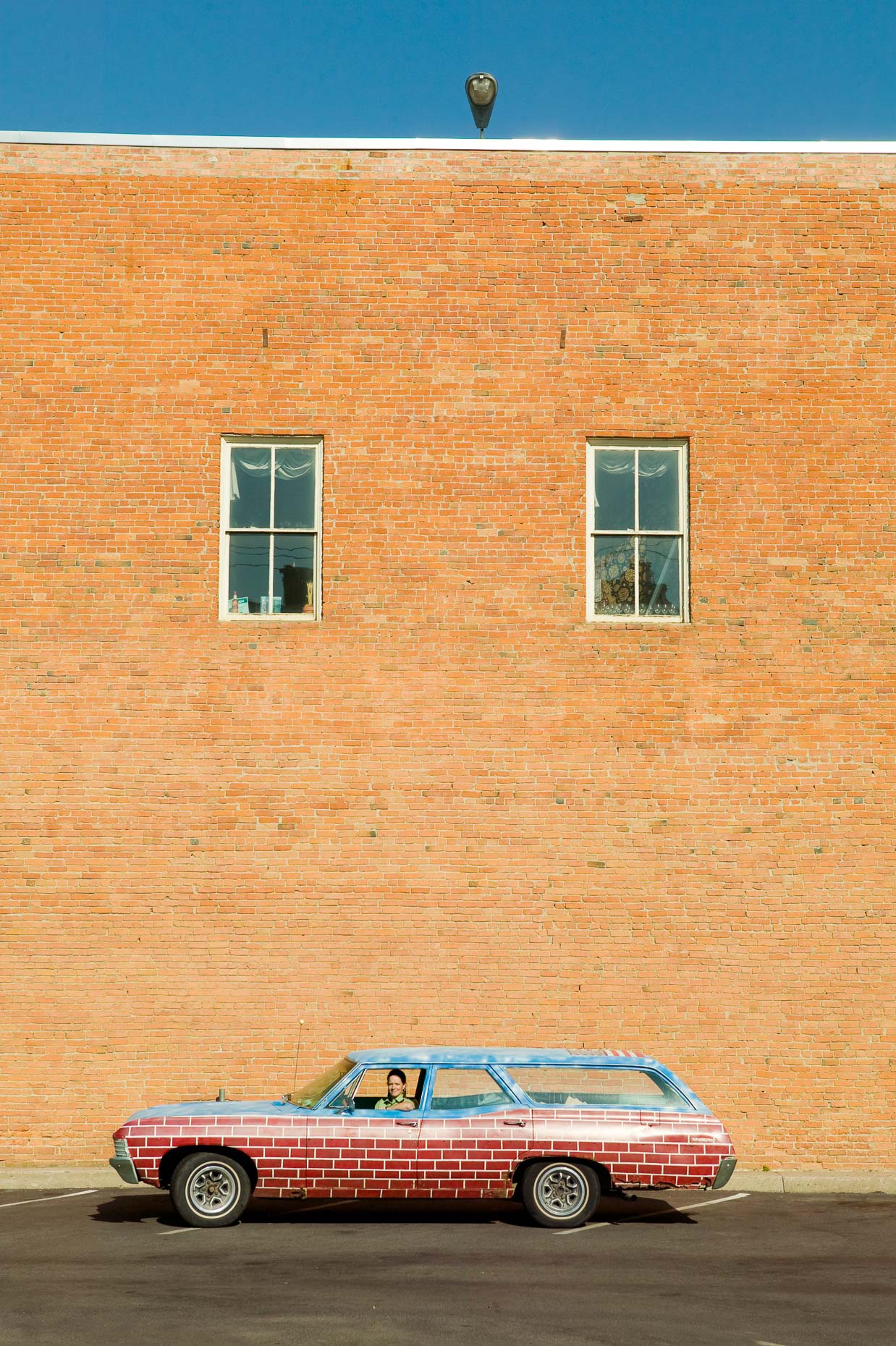 Portrait of woman in station wagon vehicle car painted with brick pattern in front of brick wall in Missoula, Montana by David Zaitz