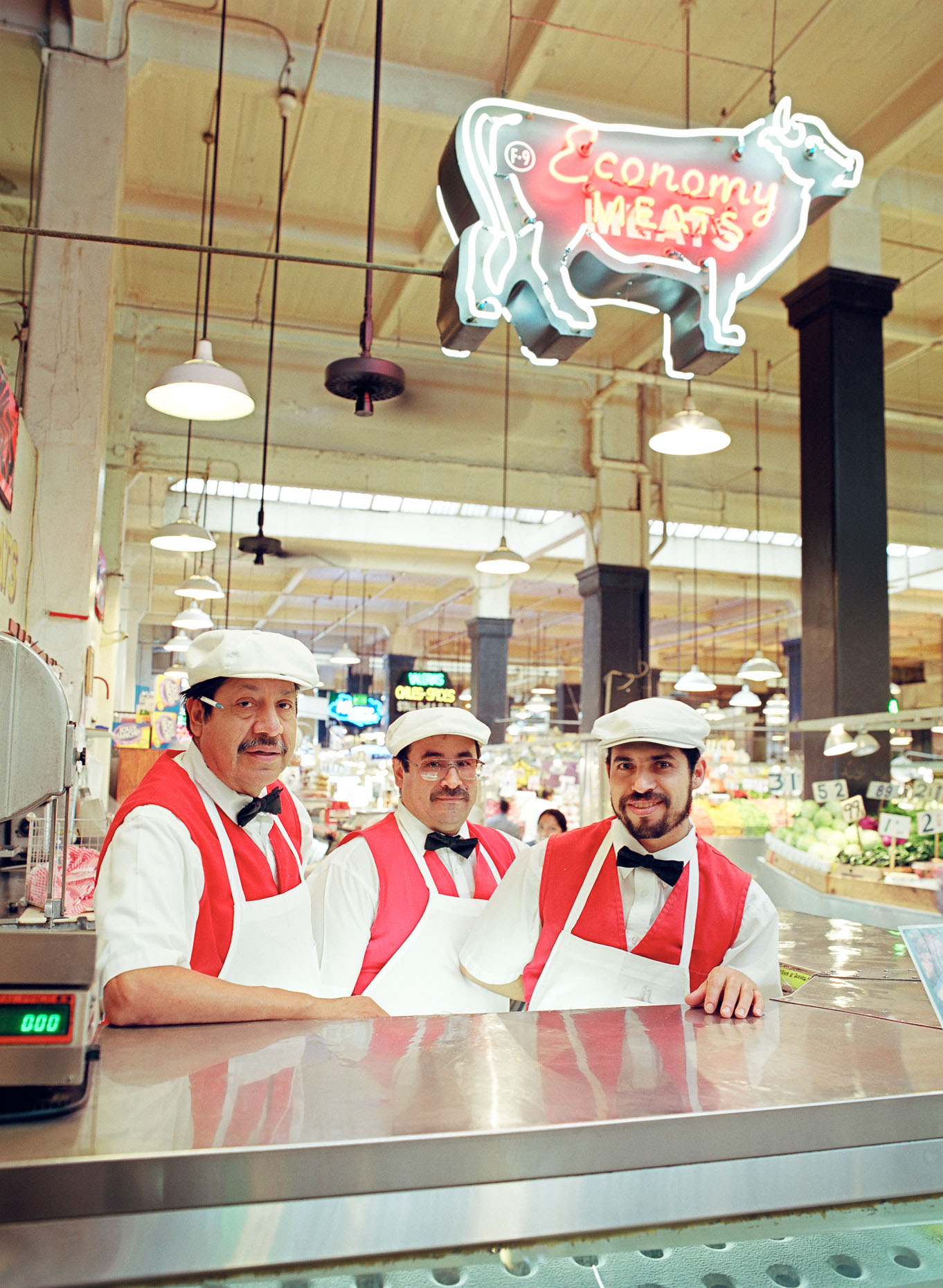 Environmental portrait of three meat butchers wearing aprons at meat market in Grand Central Market in Los Angeles, California with neon ECONOMY MEATS sign overhead.