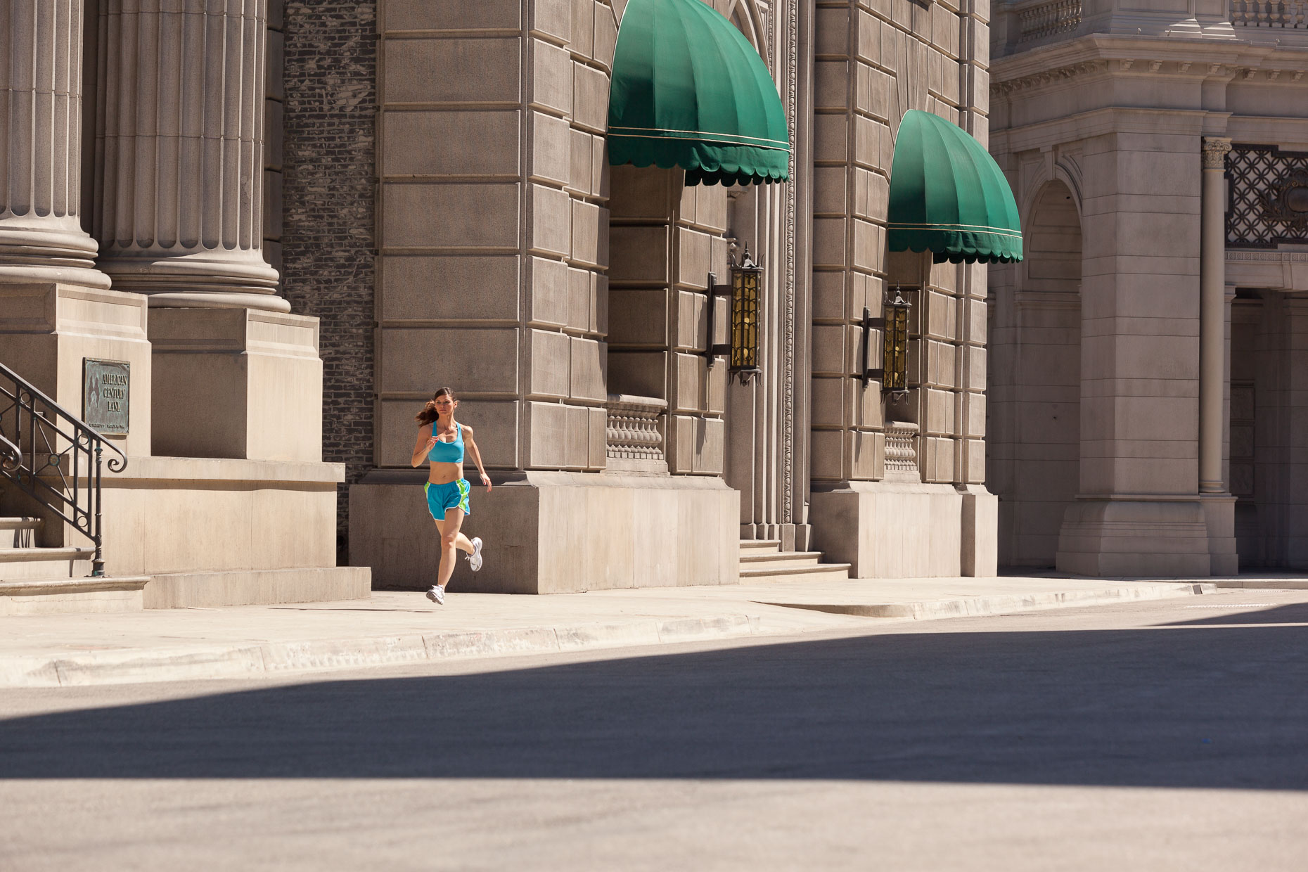 Woman in blue running clothing runs on city street with buildings in background. Fitness. David Zaitz Photography.