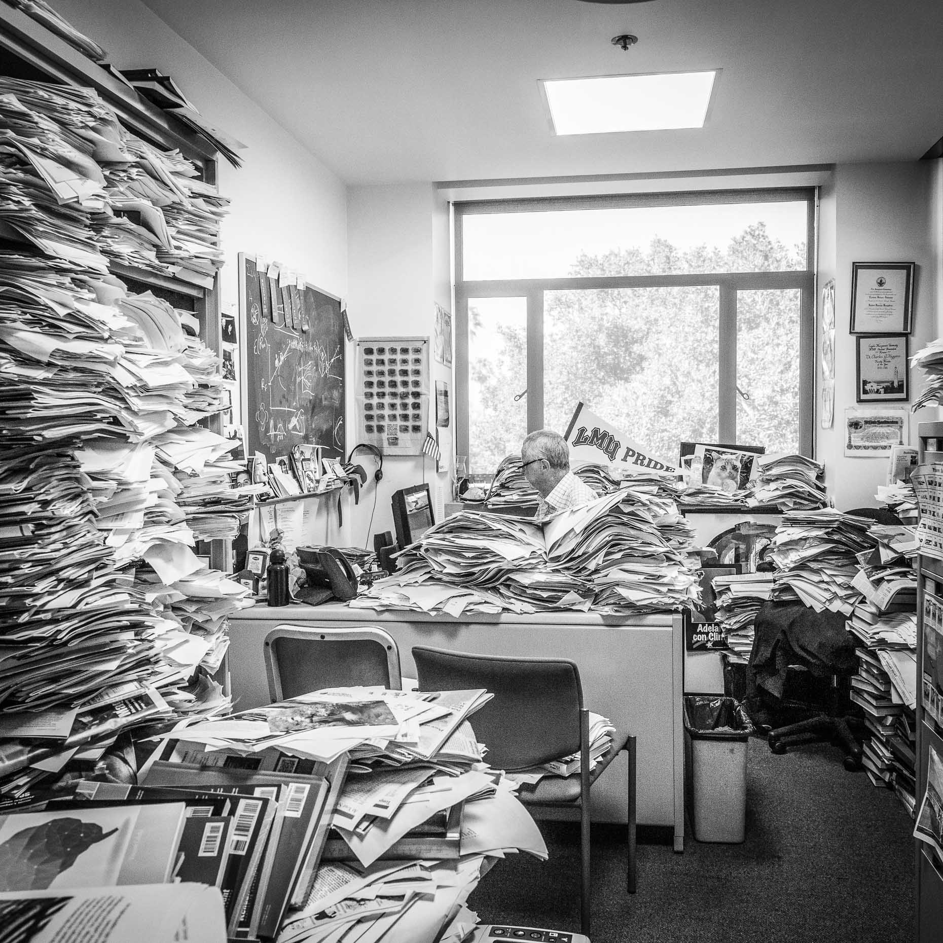 Black and white shot of man in office with many stacks of paper and folders by David Zaitz