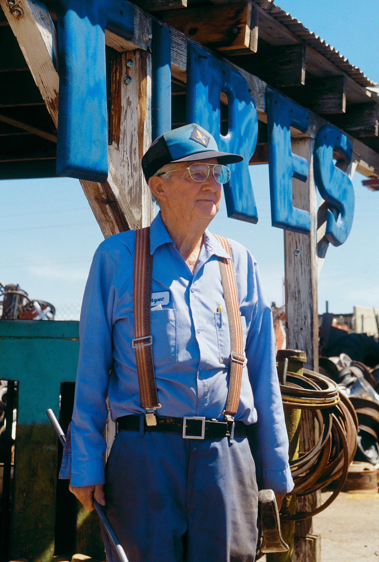 Environmental portrait of man wearing suspenders and blue uniform standing in front of TIRES sign at a tire shop by David Zaitz