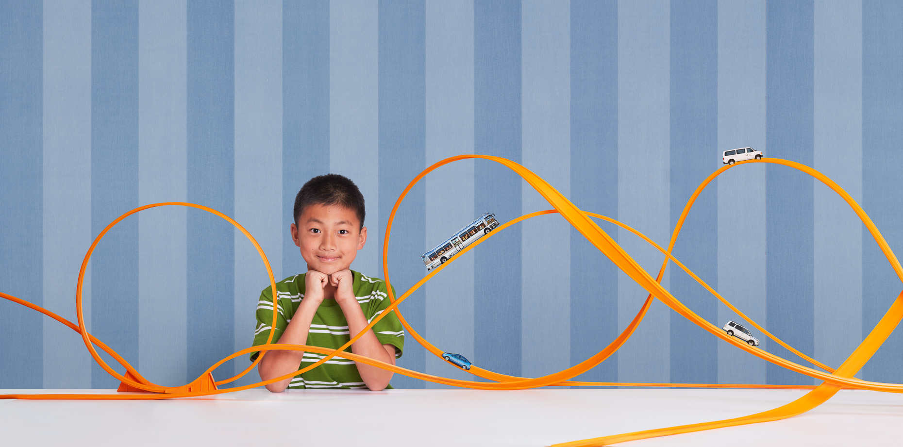 Portrait of Asian boy proud of his complex Hot Wheels toy car track setup in front of blue striped wallpaper by David Zaitz
