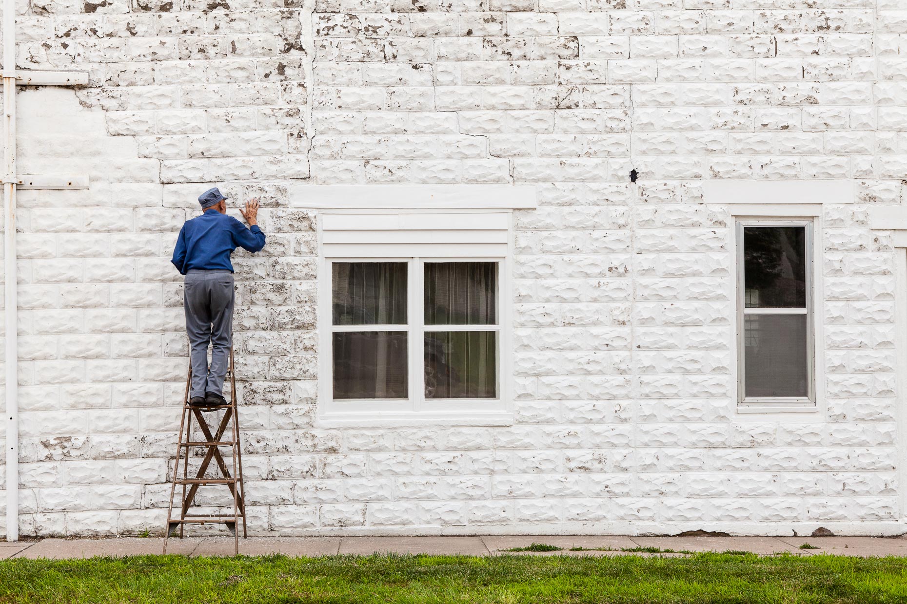 Old man standing on ladder repairs cracks in white brick wall by David Zaitz