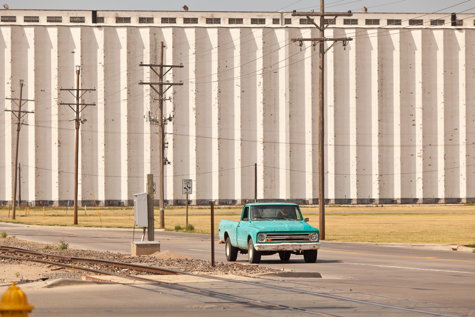 Green pickup truck driving on road with grain elevators in background by David Zaitz