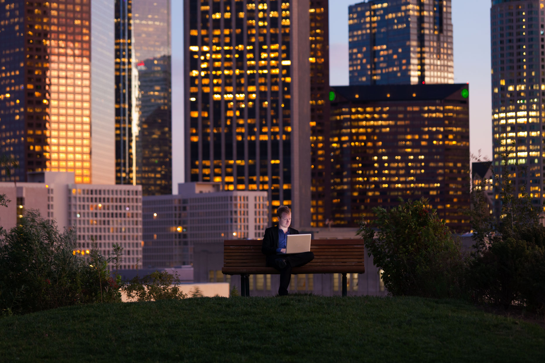 Businessman working on laptop while sitting on park bench with Los Angeles skyline in background at dusk by David Zaitz.