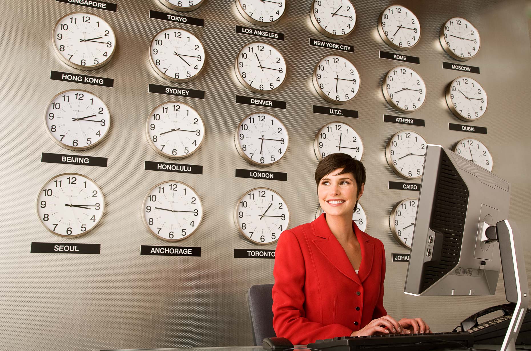 Woman working on computer in front of wall with many clocks with different international time zones by David Zaitz
