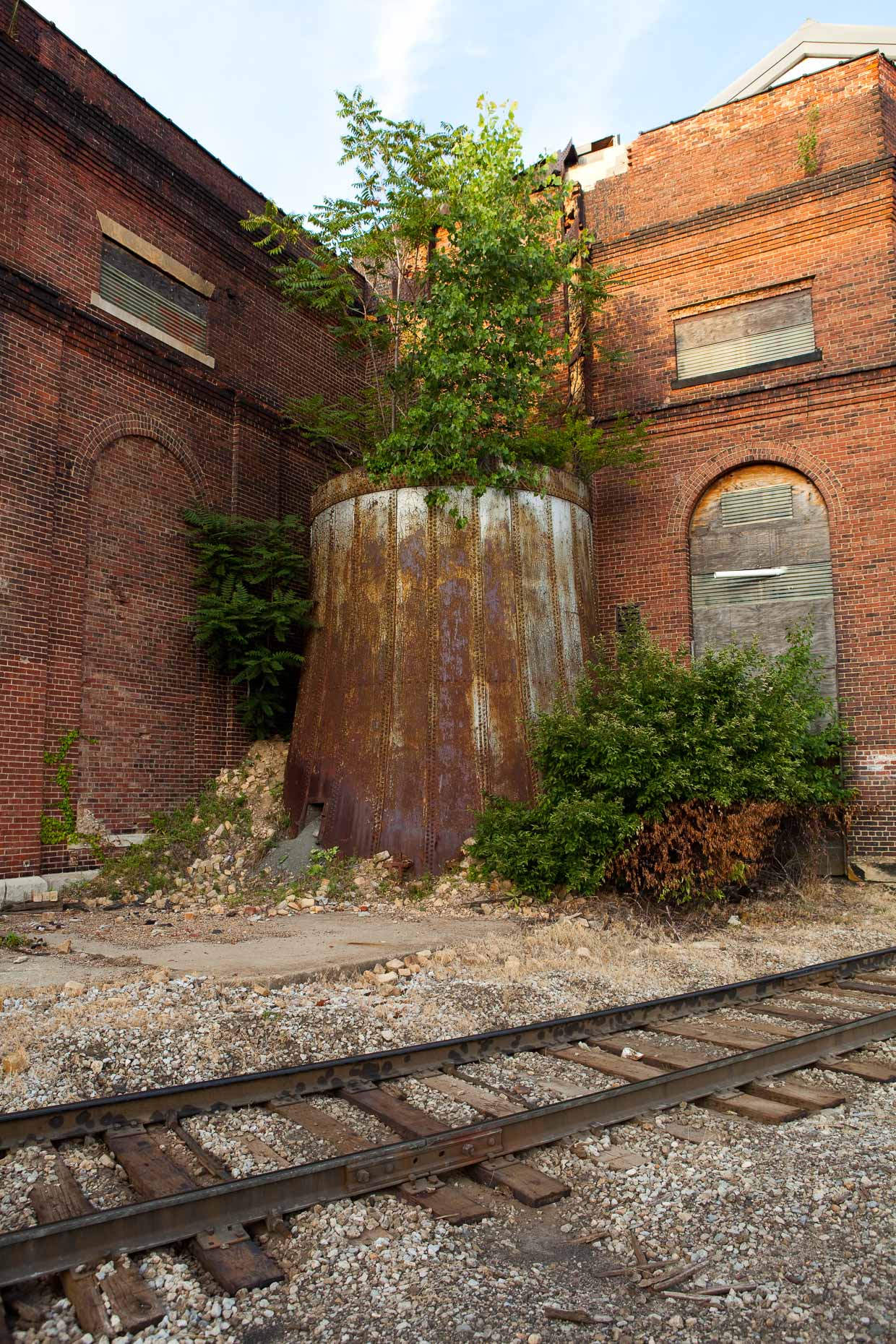 metal tank with trees growing in it next to brick factory building by David Zaitz