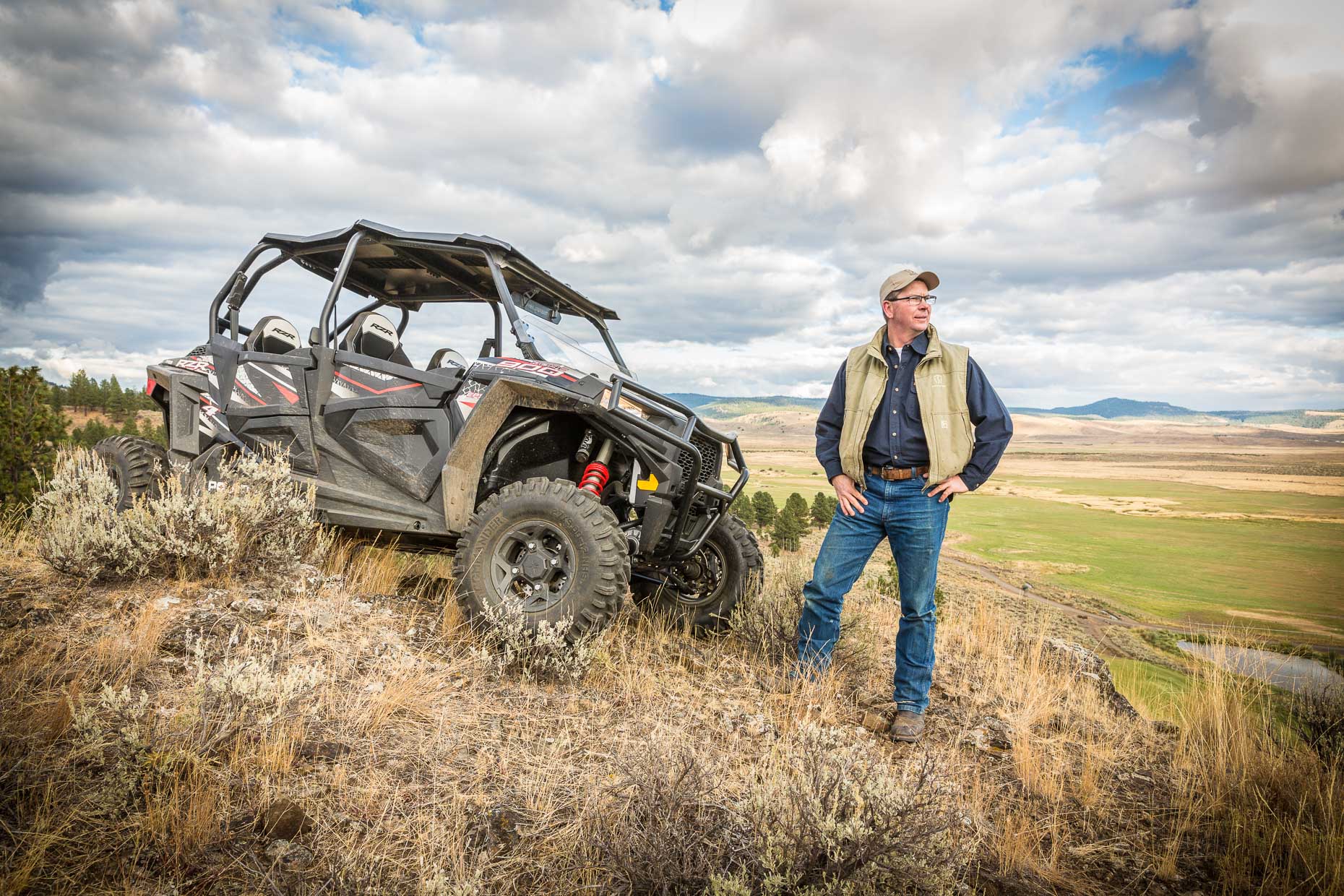 Environmental portrait of man standing next to ATV on top of hill in Seneca, Oregon.